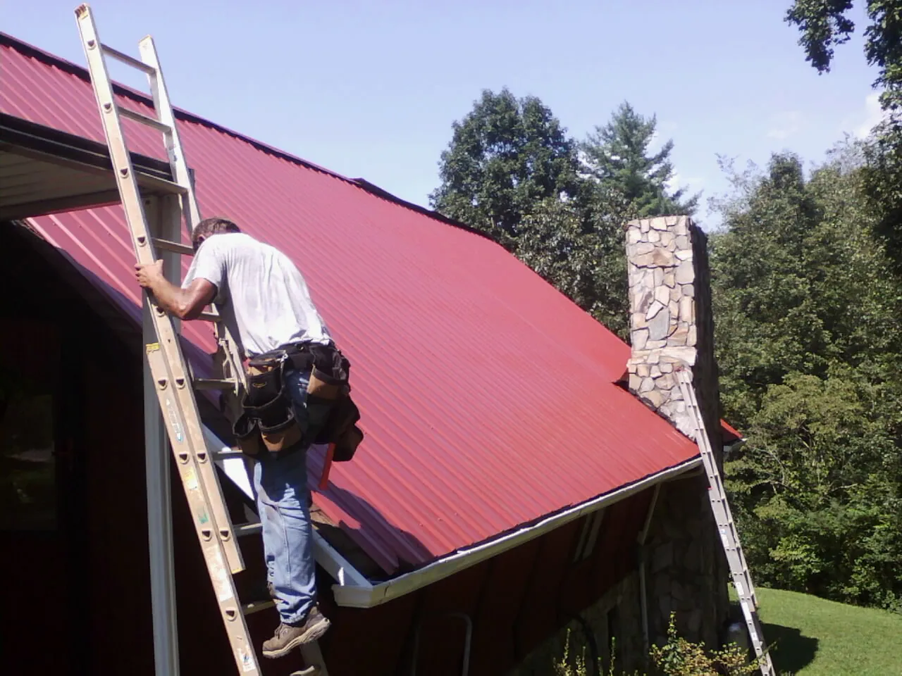Roofer on a ladder beside a red metal roof and stone chimney — Soffit & Fascia Repair by Roofing Brothers in Clear Spring